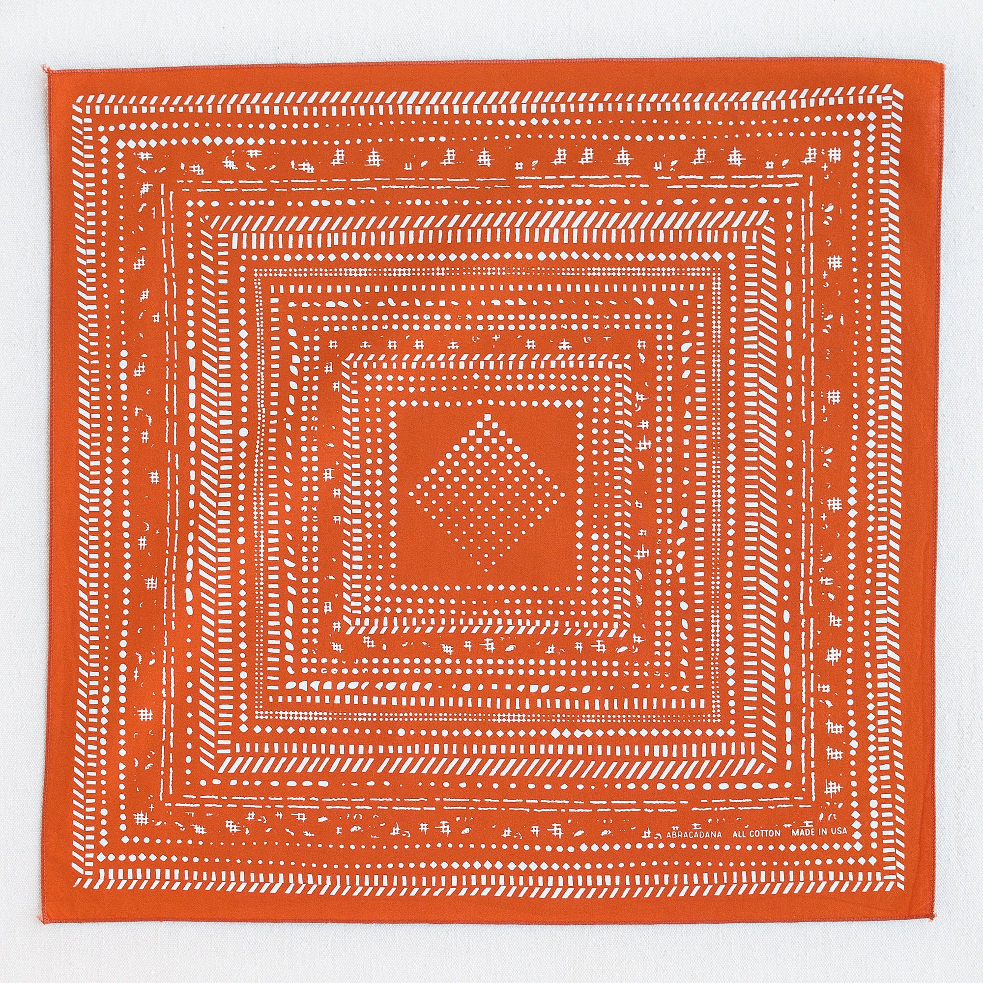 Burnt orange bandana with white print. Pattern loosely arranged squares within squares. Beginning small in the center and extending to the edge.