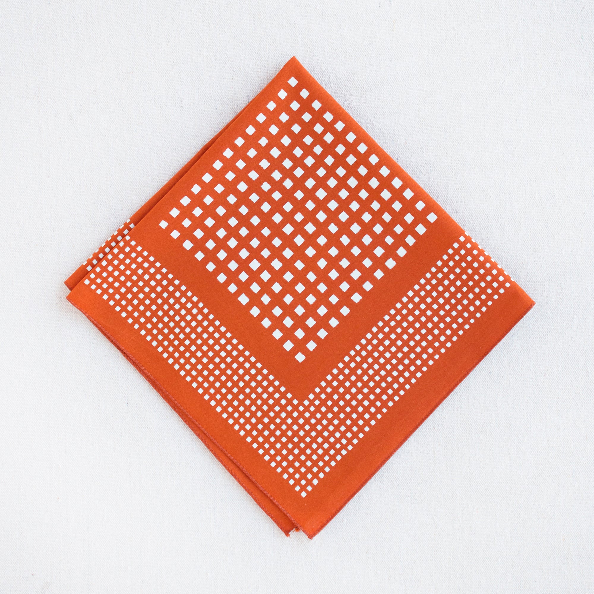 Burnt orange bandana with a white geometric pattern composed of squares. Folded into quarters. By Abracadana