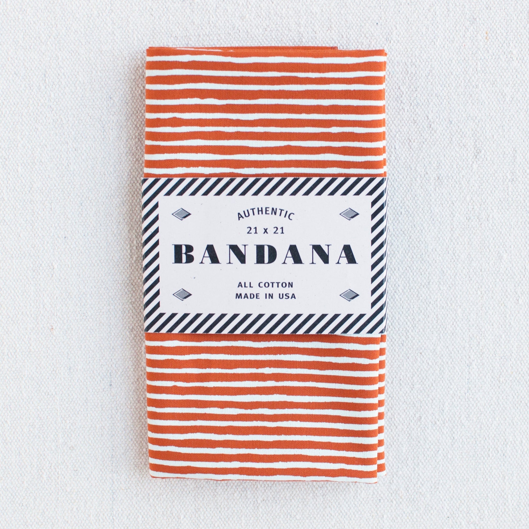 Burnt orange bandana with white striped screen print, folded and packaged. 