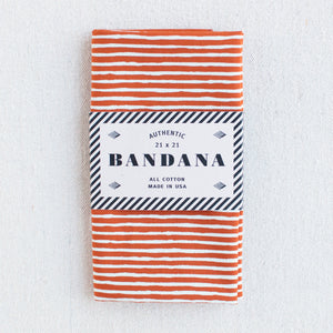Burnt orange bandana with white striped screen print, folded and packaged. 