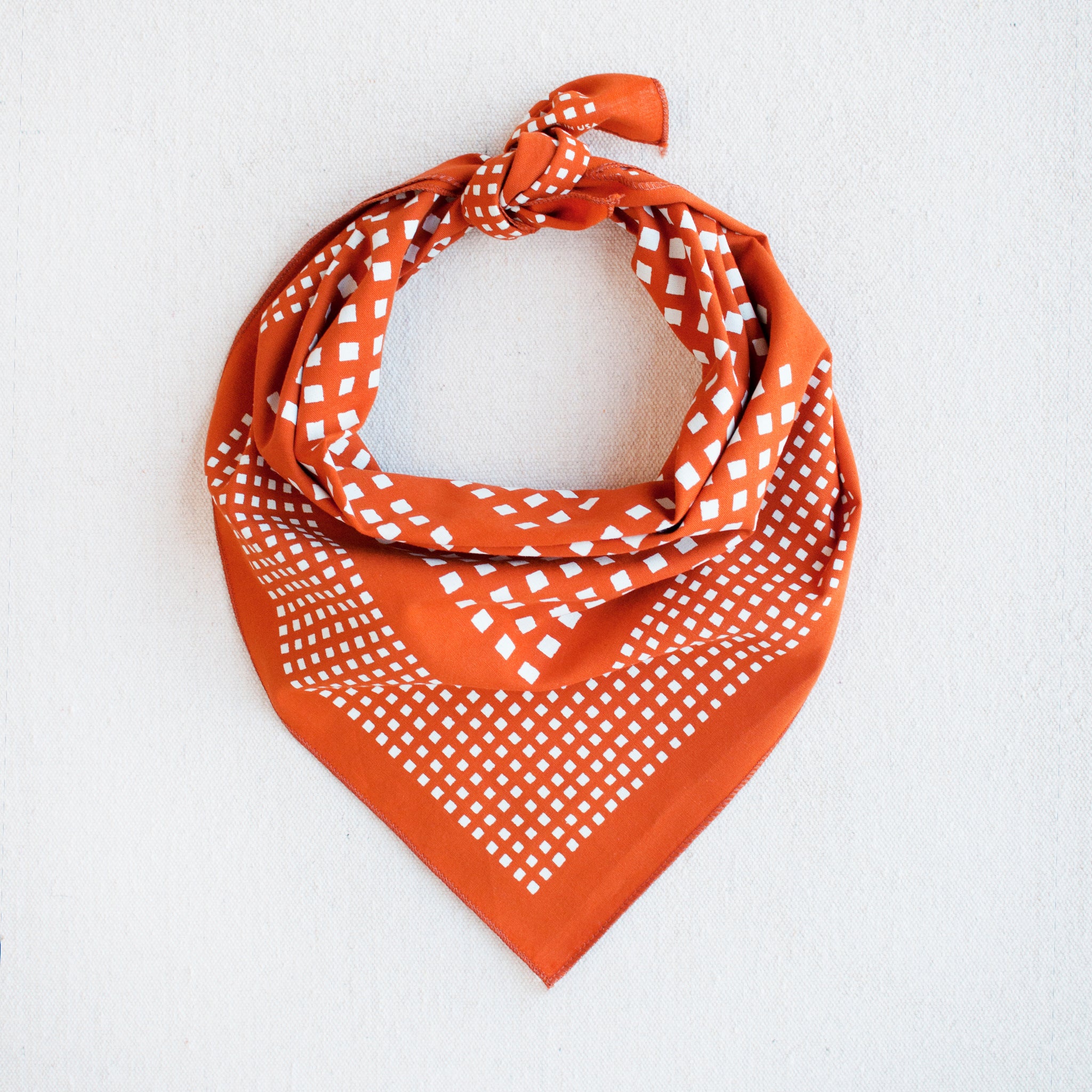 Burnt orange bandana with a white print of a square motif by Abracadana, styled as a triangle scarf.
