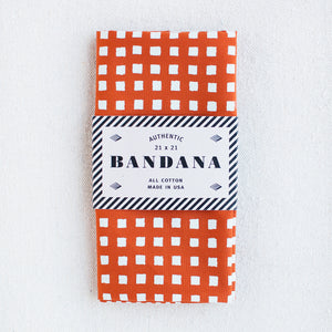 Burnt orange bandana with a white print of small squares by Abracadana, shown packaged